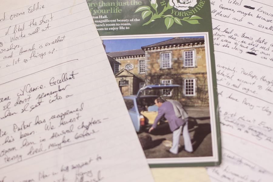 Close up of notebook paper with cursive writing in pen on it. In between two sheets is a green pamphlet and a photo of a man getting stuff out of a car in front of a dorm building.