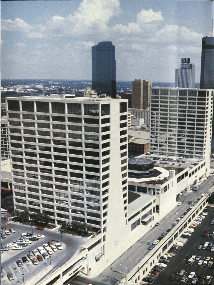 Aerial view of a downtown. There are two white buildings with several stories. Cars are seen on the bottom right of the pictures with cars driving by on the street.