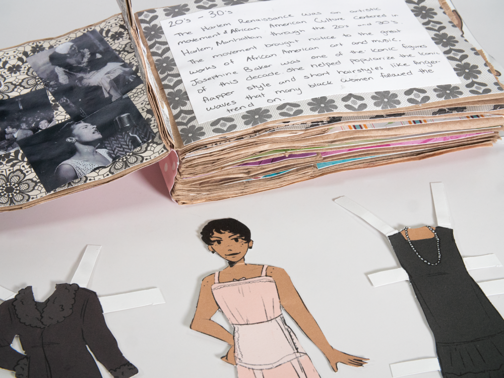 An open book made of brown paper bags, with images of Black women on the left page and text on the right. A paper doll of a Black woman sits below with an outfit on either side of her.
