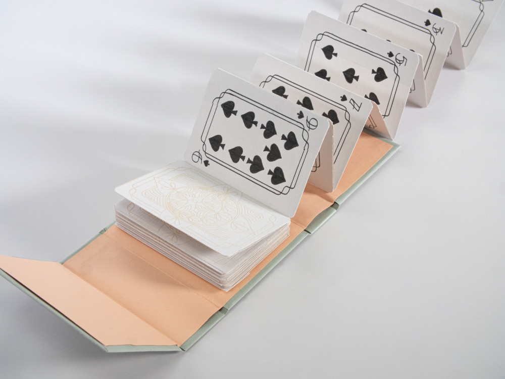 Accordion folded pages with hand drawn playing cards on one side, and an etched design on the other. They fold out from a a light pink and green case.
