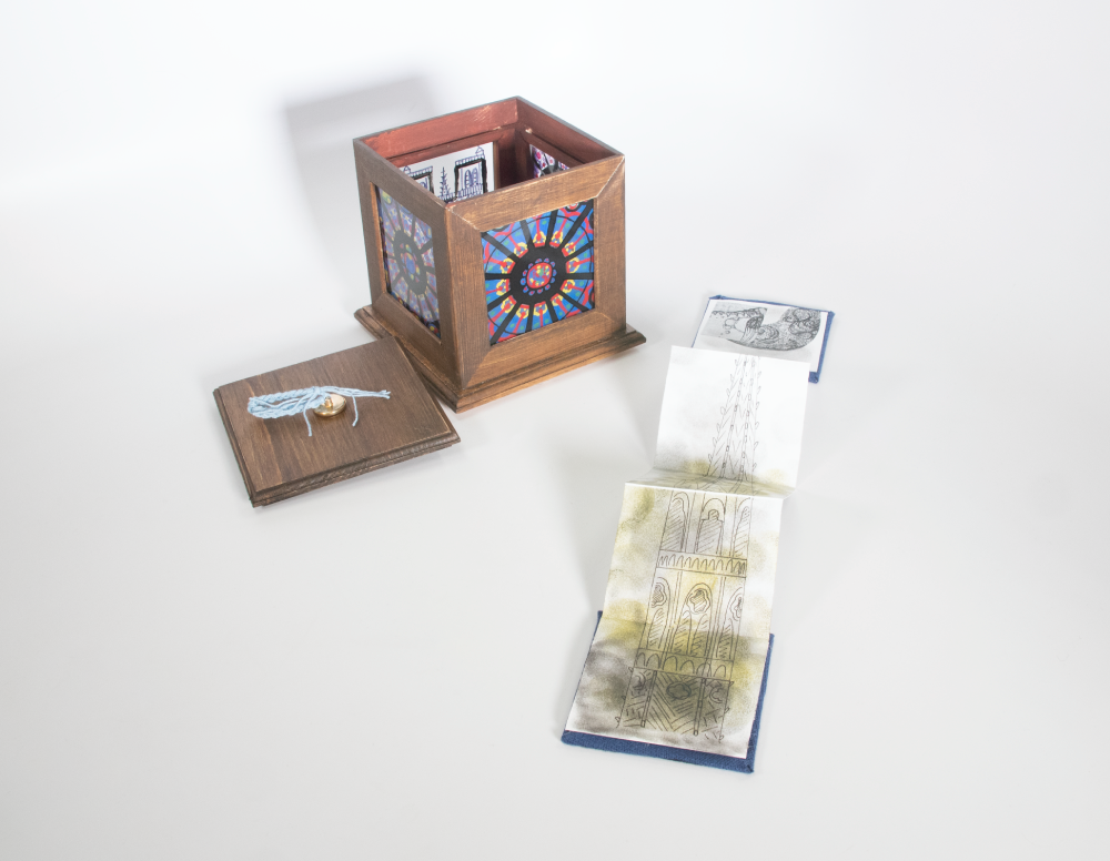 A wood cube with painted panes of glass on each side to look like stained glass. An accordion fold book is laid out next to the box with a line drawing resembling the spire of Notre Dame Cathedral.