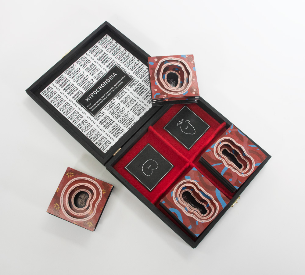 A shallow black wooden box lays open, with repeated text in the lid reading Have I Gone Mad? and a black rectangle with text at center that reads Hypochondria with the definition below. The lower portion is divided into four sections and lined with red cloth. Each section has a different three dimensional pop up paper cube.