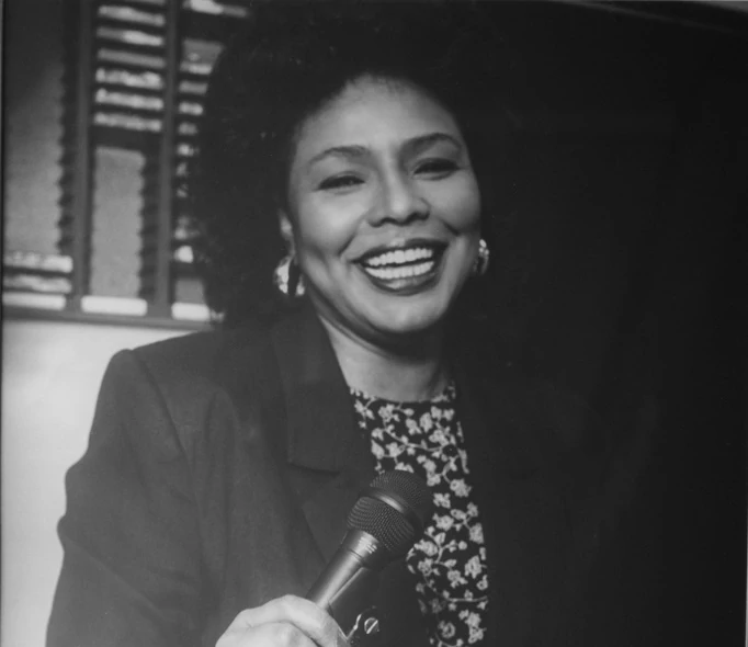 Black and white close up photograph of Iola Johnson holding a microphone and smiling.