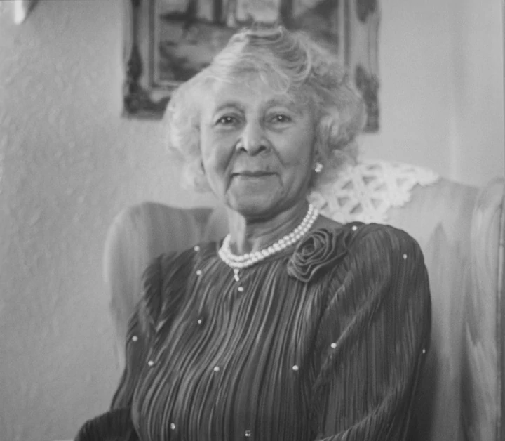 Black and white photograph of Ollie McMillian-Mason seated in a chair with doilies on the arms and back. She wears a long sleeve dress and pearls.