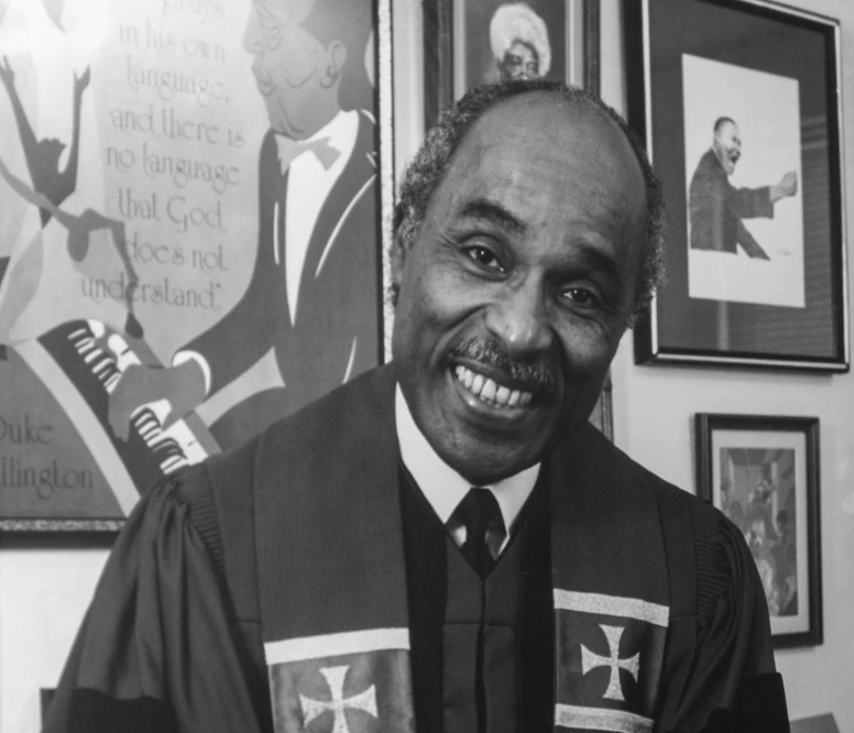 Black and white photograph of Rev. Zan W. Holmes Jr. standing in front of a wall of art and photographs. He wears robes and a sash with crosses on either side of his chest.
