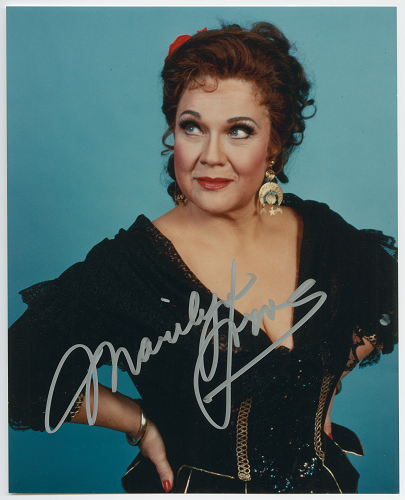 Photo of a woman with something red in her hair and in a black dress with a corset. A white signature is at the bottom of the photo.