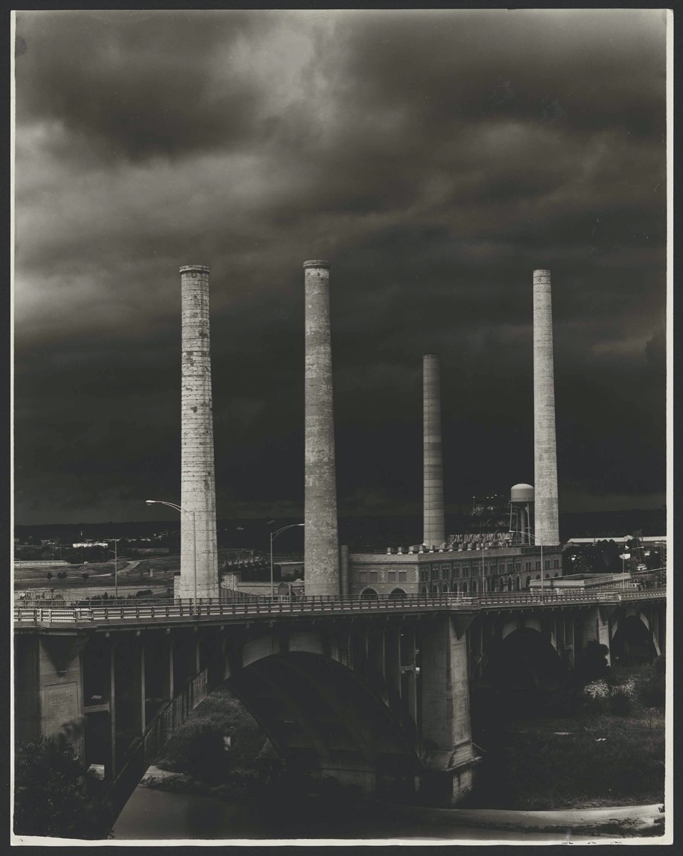 Black and white photo of a power plant, four white towers in the air. It is seen from a distance.