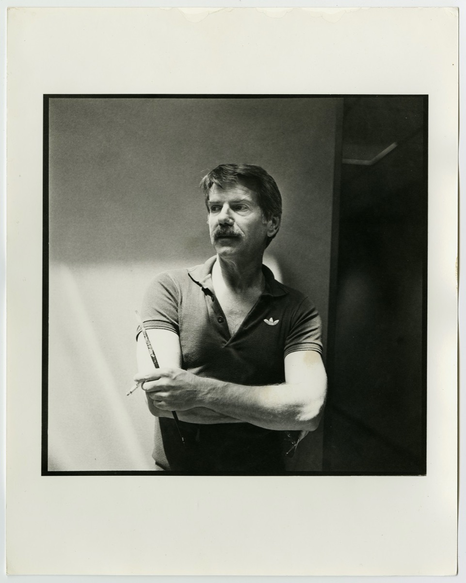 Photo of a man in an unbuttoned polo shirt and a moustache. His arms are across each other.