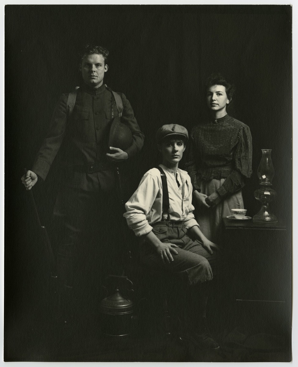 Three people posing for a photo. On the left a man stands with a rifle propped on the floor, a helmet in his arm and straps on his shoulders. In the middle is young man with suspenders and cap, sitting on a chair. On the right is a woman in longsleeves and a skirt.