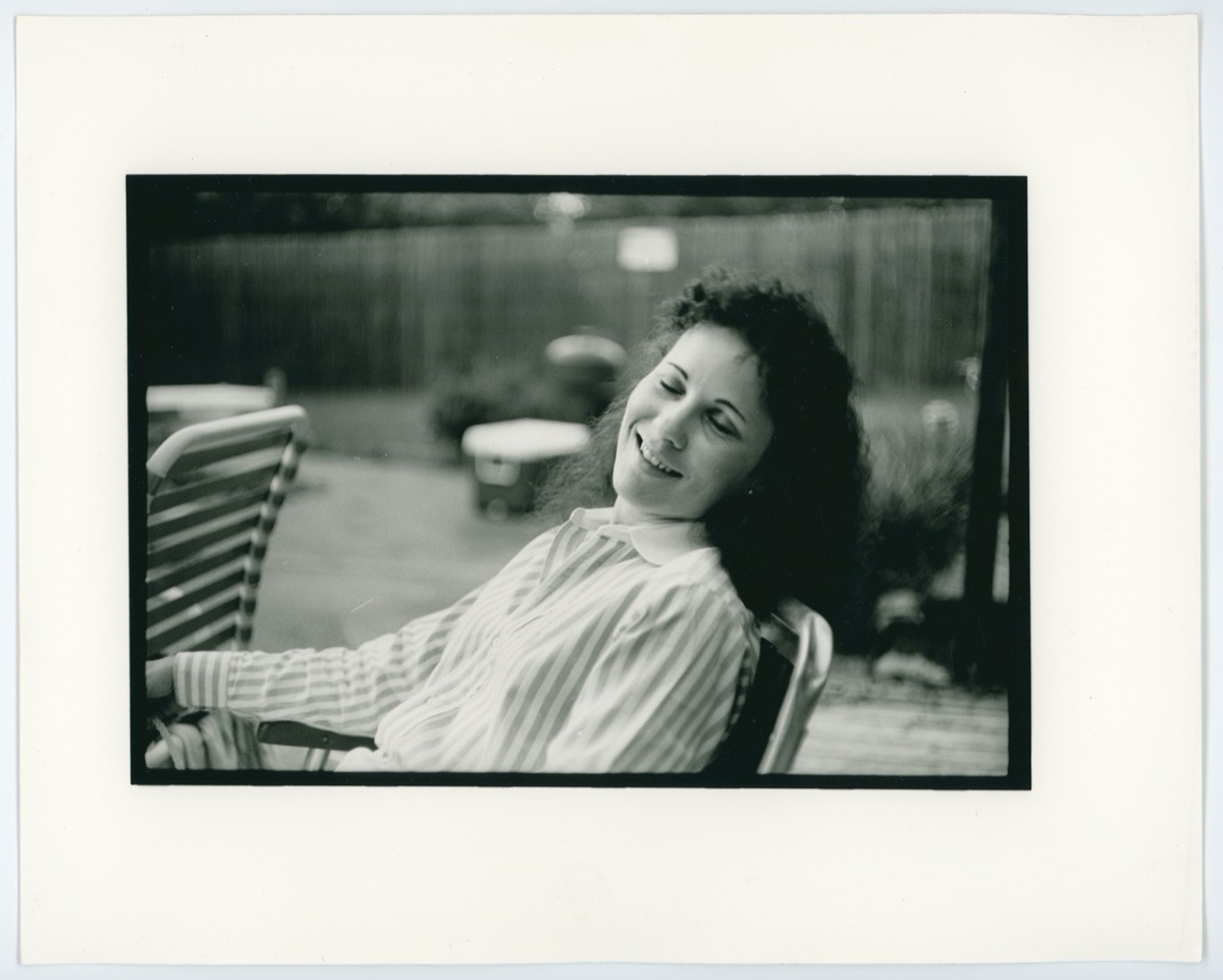 A young woman in a white striped shirt sits on a chair outside, leaning backwards with her eyes closed.