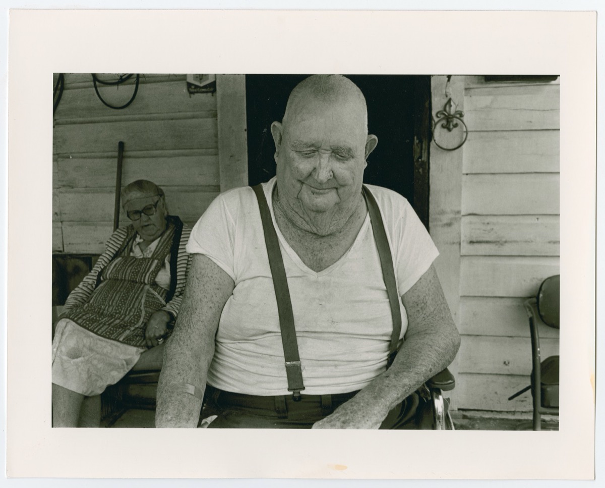 Bald old man in a white shirt and suspenders, looking down and sitting in a chair. Behind him on the left side of the photo is an old woman with an apron on, sitting on a chair against the wall of a house.