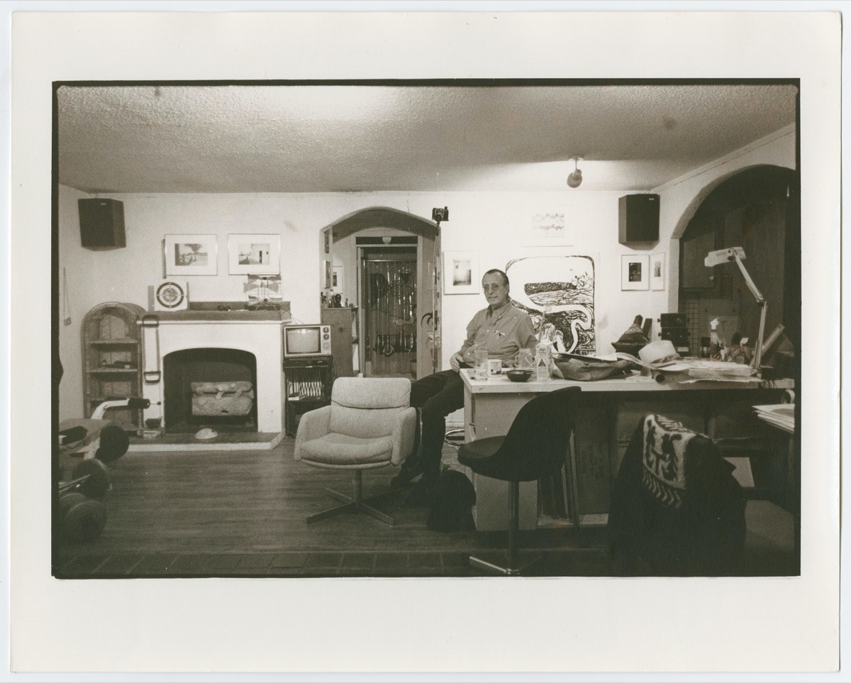 Man sits on a chair, a messy desk beside him. To his left iis a doorway, and a fireplace with two framed photos above it.