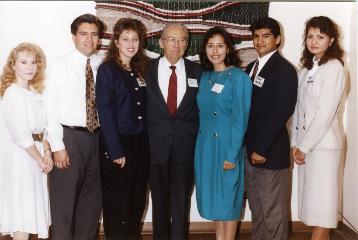Group of 3 men and 4 women. The one in the middle is an old man in a black suit and red tie. There are three people on each side of him. On the right, beginning from the left, is a woman in a light blue dress, a man in a black suit and brown pants, and a woman in a white dress. To the left of the man in the middle (from right to left) is a woman in a navy suit, a man in a white dressy shirt and tie, and lastly a woman in a white dress.