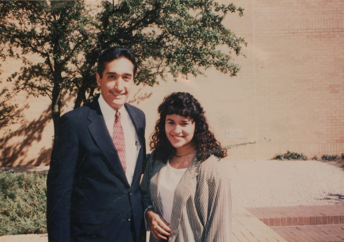 A man in a suit poses arm in arm with a woman to his right in the picture. Behind them is a tree and a brick wall.