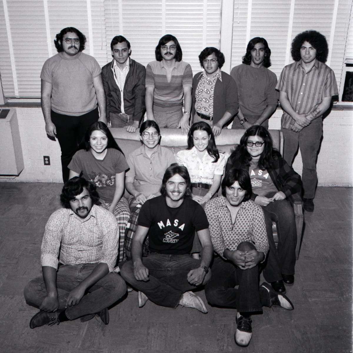 Group of 13 people. The first row is 3 men sitting down, the middle row is 4 women sitting on a couch behind them. Behind the couch is a row of 5 men and 1 woman standing up.