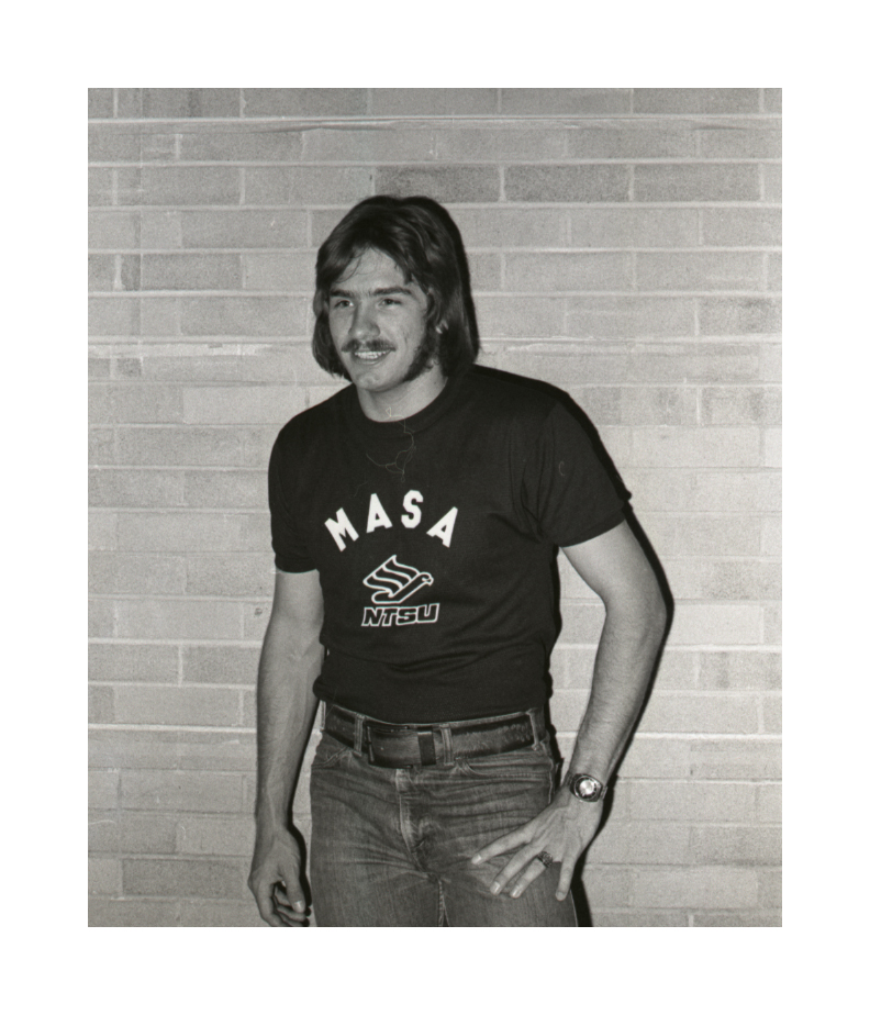 A man stands in front of a brick wall, posing with his left arm on his left. He has a moustache and wears a shirt with the letters MASA and NTSU on it.