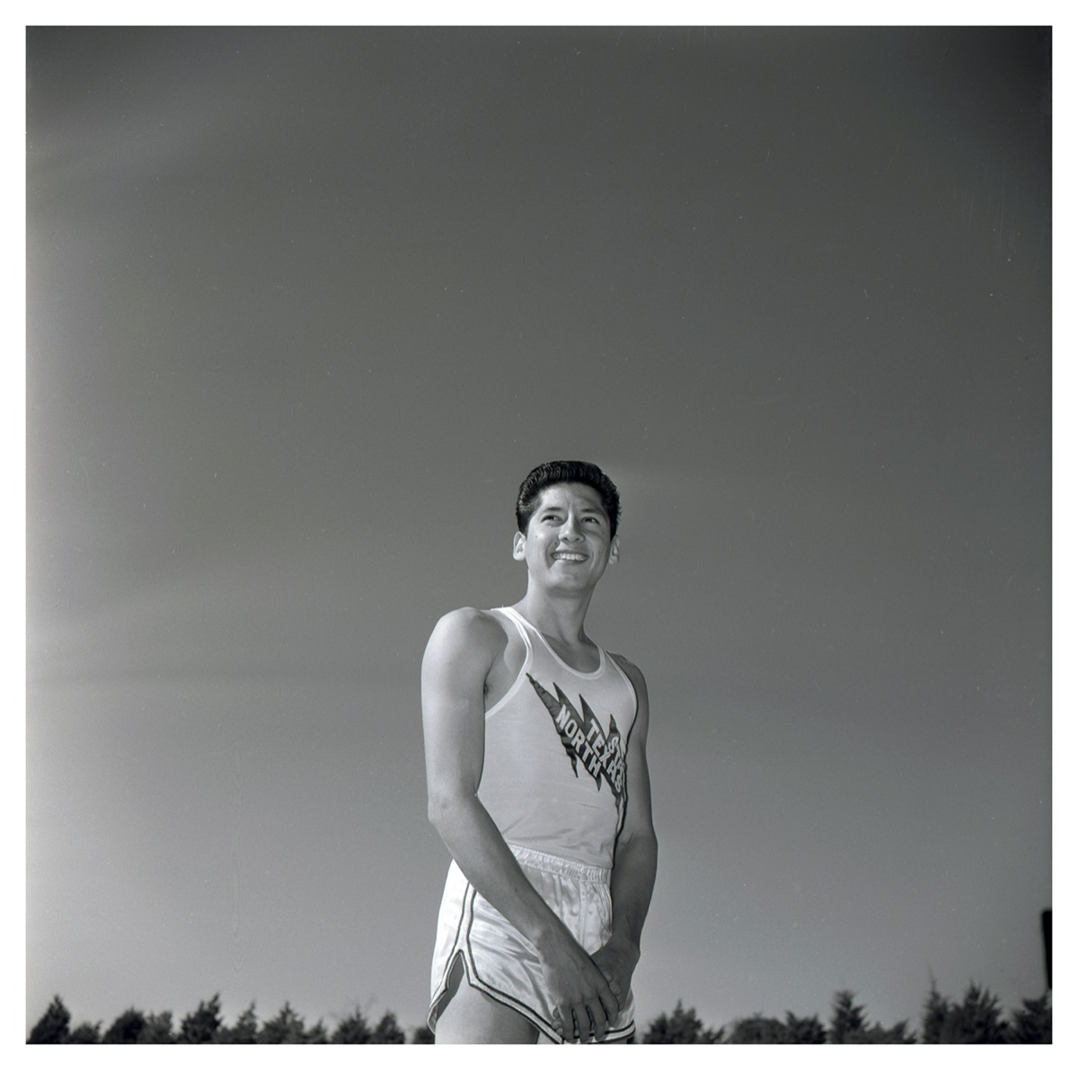 Black and white photo of a man in short white shirts and a white tank top with the words North Texas on it. He is seen from the upper leg up with the clear sky seen behind him.