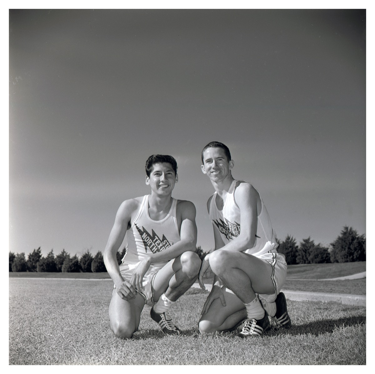 Black and white photo. Two men kneel down posing side by side each other. The both wear the same  white shorts and white tank tops. They are kneeling on the grass, with sky  and an outline of trees behind them in the distance.