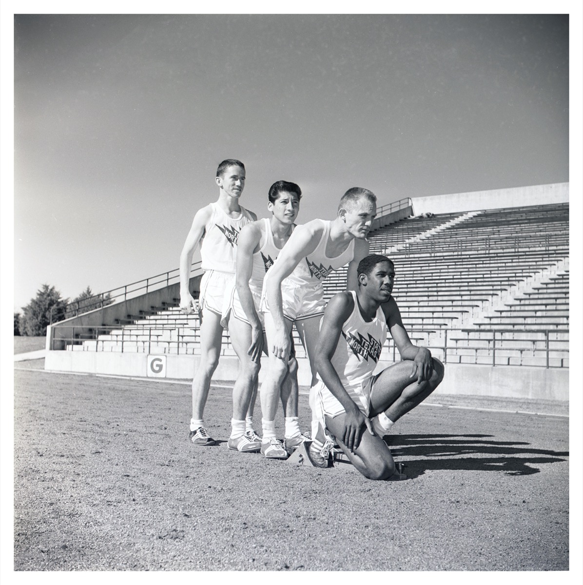 Black and white photo where 4 men pose in line behind each other. They all wear the same white shorts and tank top. The bleachers are seen behind them.