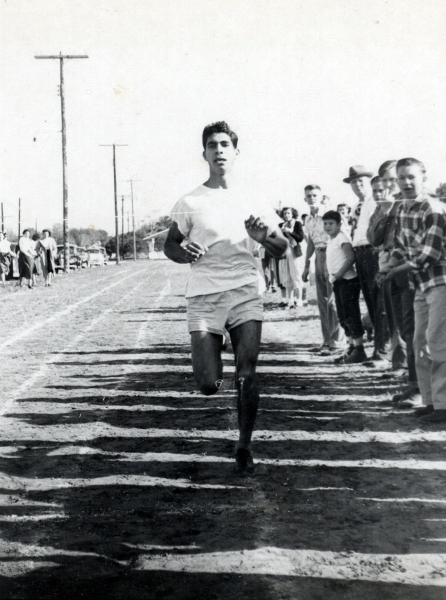 Black and white photograph of a man in a white shirt and shorts runs on a track. To his right is a row of people watching him.