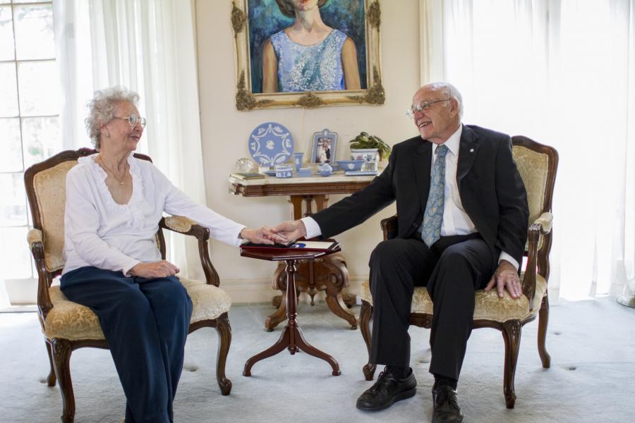 Una anciana se sienta en una silla a la izquierda de la foto, mientras que un anciano se sienta en una silla en el lado derecho de la foto. Se sonríen mientras se cogen de la mano. Hay una pequeña mesa entre sus sillas, parte de un cuadro que se ve en la pared detrás de ellos.