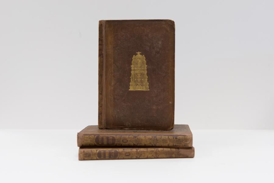 One book is propped on top of a stack of two books. They are all a brown color. The book on top has golden clock on it, but is otherwise plain.