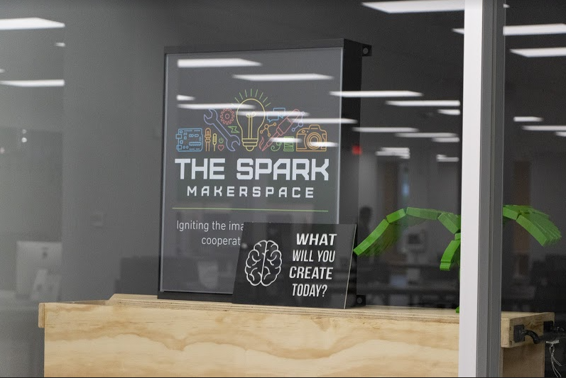 Sign behind a window that says The Spark Makerspace, with a line graphic of a lightbulb, camera and various tools across the top. Another sign has a brain and reads What will you create today?