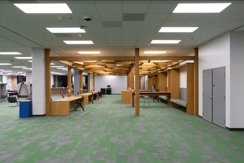 Fotografía en color de una pérgola de madera con formas geométricas creadas a lo largo del techo. Debajo hay una serie de escritorios de madera con sillas y bancos acolchados a lo largo de la pared.