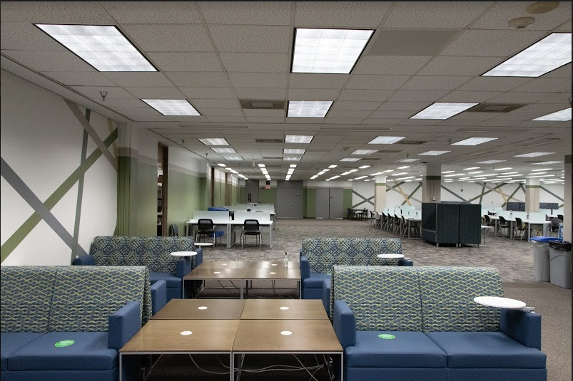 Color photograph of large open floor, with various furniture, including small blue and green patterned sofas with tables in between them, desks with partitions, and large cube seats. The walls are white with diagonal lines colored blue, green, and beige, going in various directions.