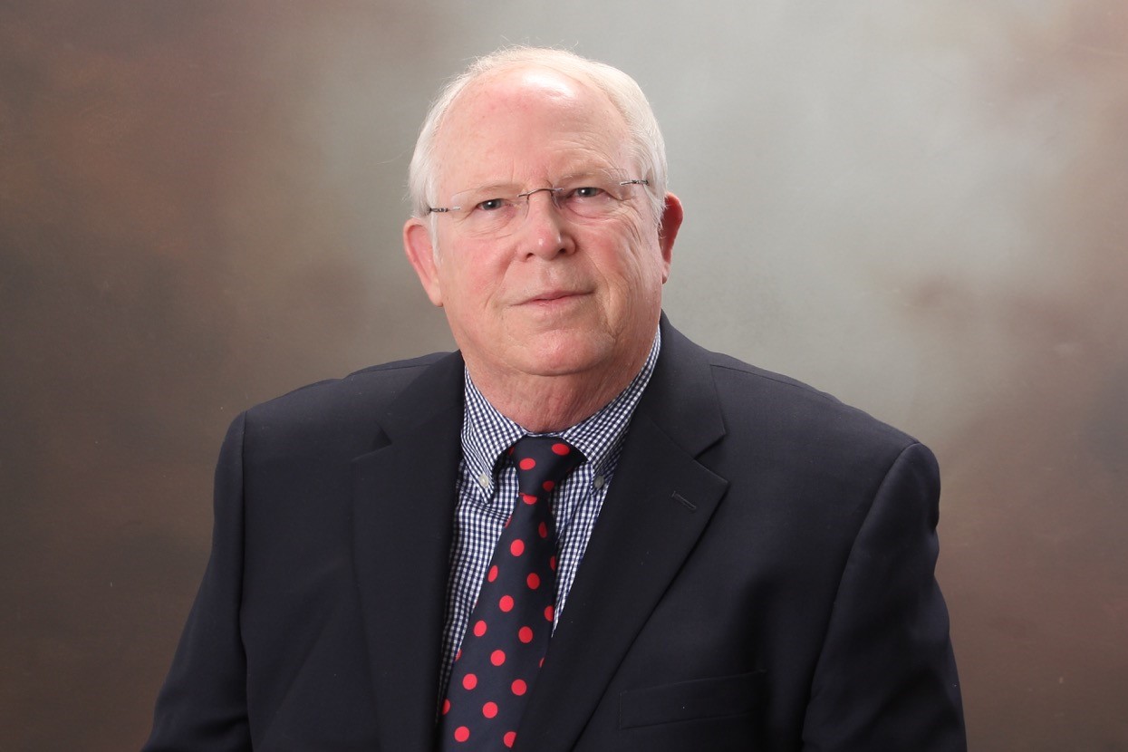 Photo of an old man with thin glasses, wearing a navy suit with a navy tie with red polka dots. He wears a blue and white plaid shirt underneath, the background is grey in color.